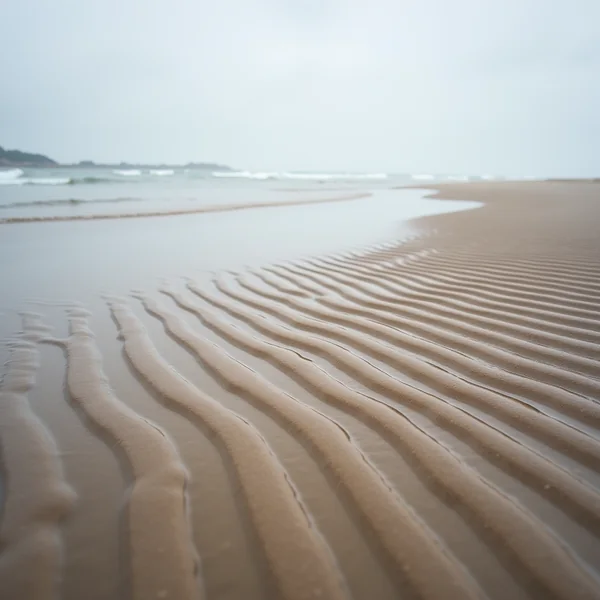 Ripples of wet sand across a wide flat beach at low tide, the sea and a faint headland beyond under an overcast sky