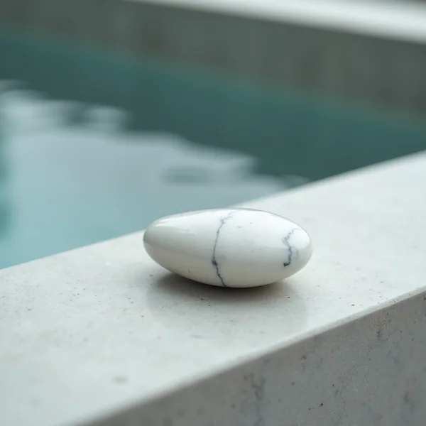 A smooth white veined marble pebble resting on a pale stone ledge, with still water out of focus behind it