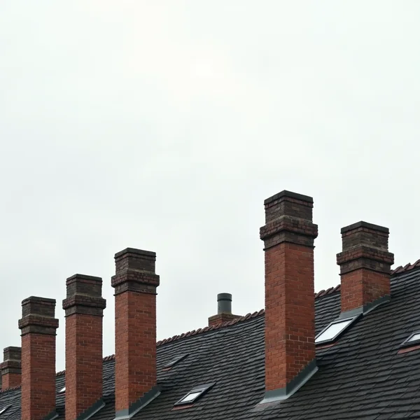 A row of tall red brick chimney stacks rising in line along the ridge of a terrace of tiled rooftops set with skylights, against a flat pale overcast sky